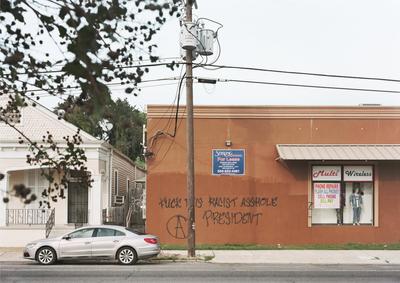 color image of street scene; white house on L with black metal railing around porch and stairs to front door; orange building on R with advertisement signs for "Multi Wireless" and two mannequins in window, as well as a blue "For Lease" sign at center, over black graffiti that reads: "Fuck this racist asshole president"; silver sedan parked on street in foreground