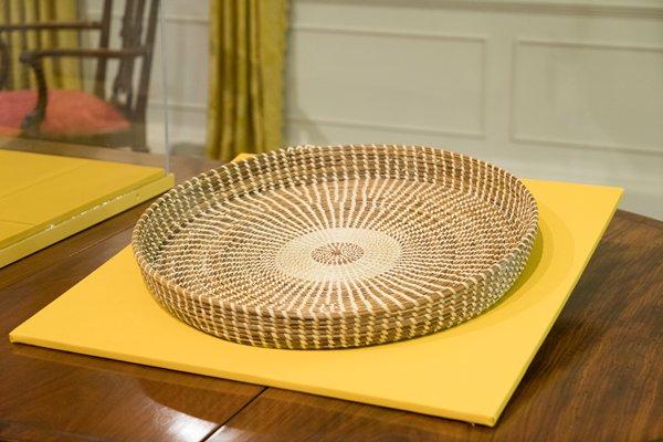 large shallow round woven circular basket; alternating woven bands of grass in light and darker shades of tan; circular pattern in center